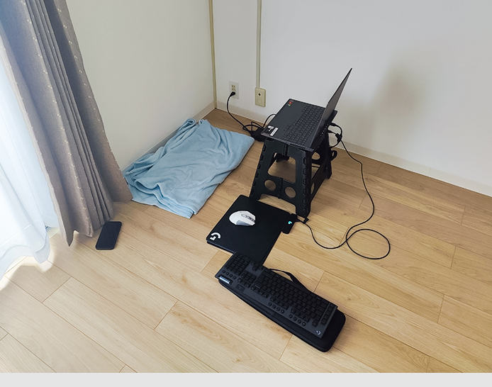 Improvisierter Arbeitsplatz: Auf dem Hellholz-Laminatboden liegt eine hellblaue Decke, als Ersatz für ein Sitzkissen. Rechts davor ein schwarzes Mousepad mit einer weißen PC-Maus und einem schwarzen Plastikhocker, auf dem ein schwarzer Laptop steht.