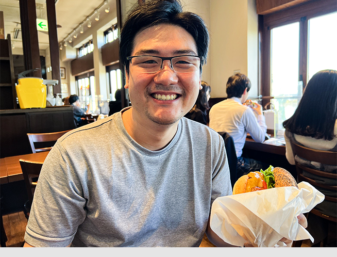 Yasuyuki sitzt in einem Restaurant, lacht glücklich und hält einen japanischen Burger in der Hand.