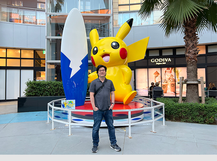 Im Hintergrund ist ein Gebäudekomplex mit vielen Fenstern. Davor eine Hecke und rechts im Bild eine Palme. Im Vordergrund zentral steht eine 3 Meter große Pikachu-Statue und Yasu posiert davor mit den Händen in der Tasche.