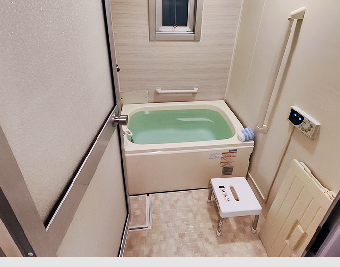Ein kleines Badezimmer mit einem weiß-beige gefliesten Boden. An der rechten Wand ist eine Heizung befestigt, ein digitales Thermostat angebracht und ein weißer Hocker steht vor der ganz hinten im Raum passend platzierte Badewanne.